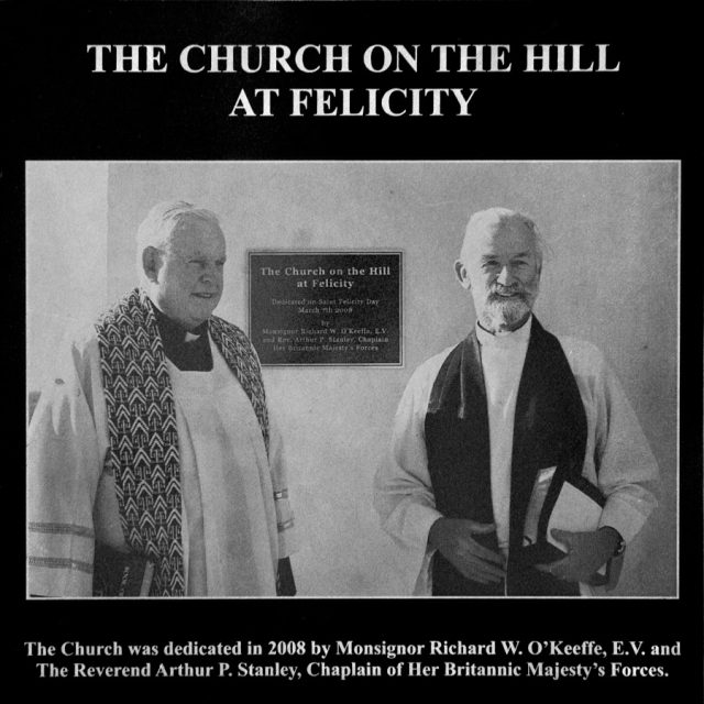 Two older men in religious attire stand in front of a plaque which reads The Church on the Hill at Felicity