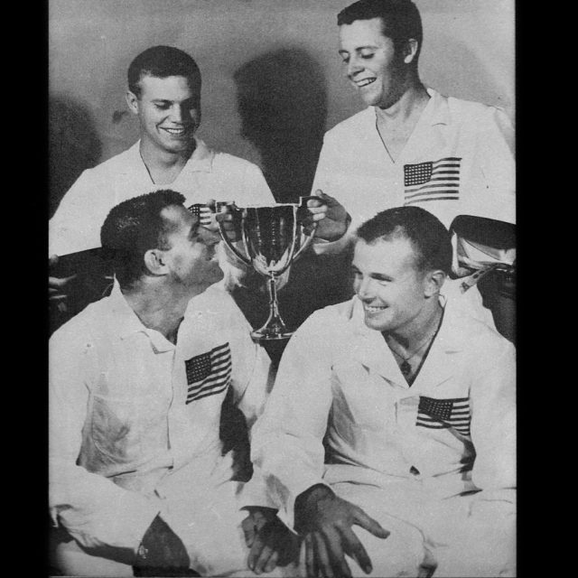 Engraving of four men in white uniforms with the American flag over their left breast smile holding a trophy.