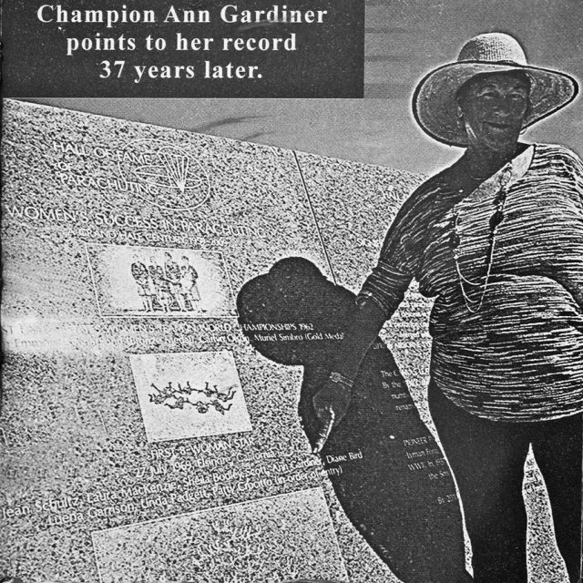 An older woman in a widebrimmed sunhat points to granite panel dedicated to the Hall of Fame of Parachuting which lists her name as a record holding female skydiver