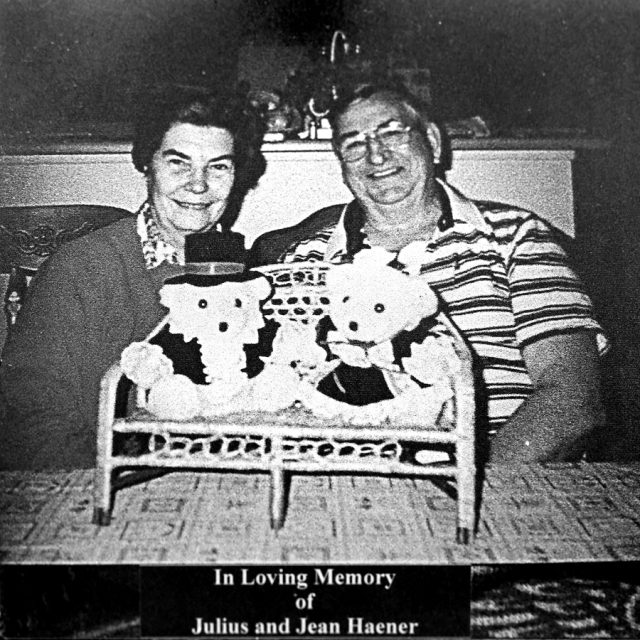 An elderly man and woman smile seated behind two teddy bears placed on a miniature wicker bench