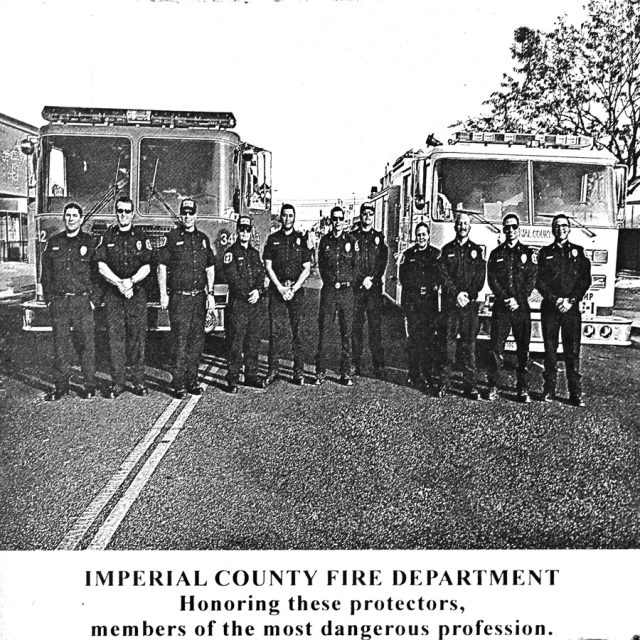 Members of the Imperial County Fire Department stand with hands folded in front of fire trucks