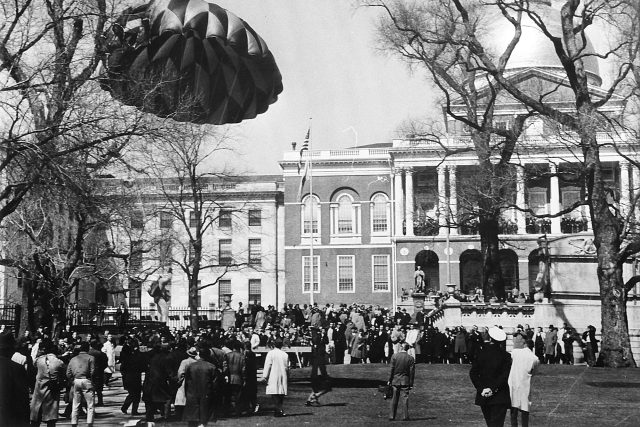 A parachutist beneath a round parachute lands in front of a grand governmental building in Boston as a crowd watches