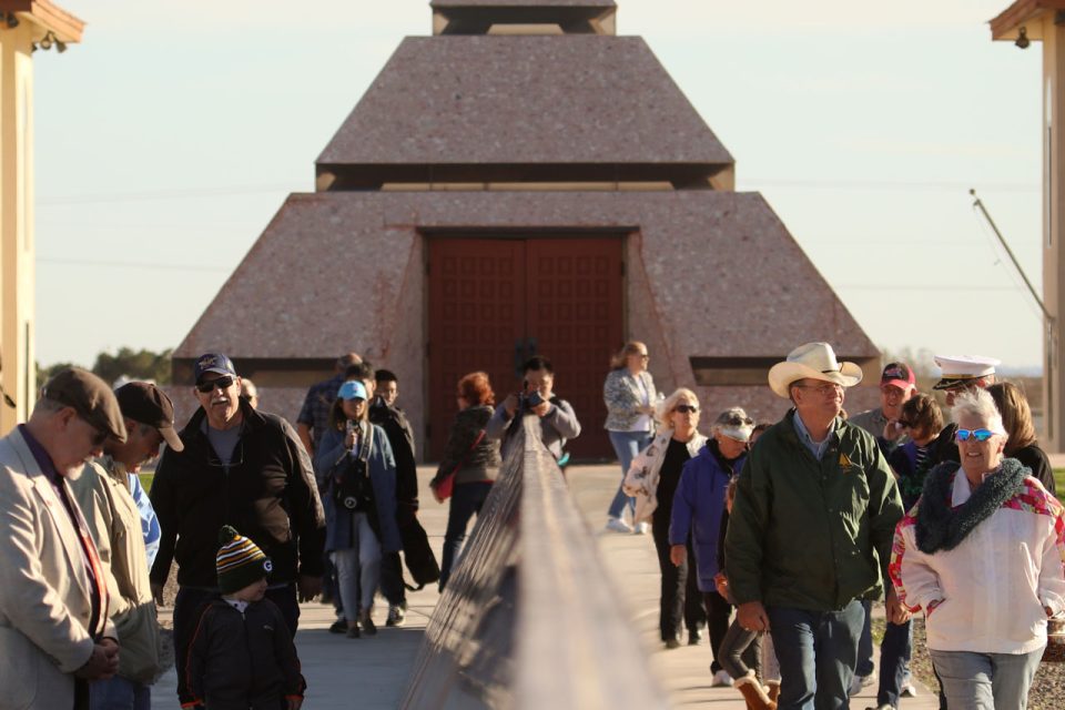 Crowd of visitors walk along granite monument at the History of Humanity in Granite with Center of the World pyramid in the background