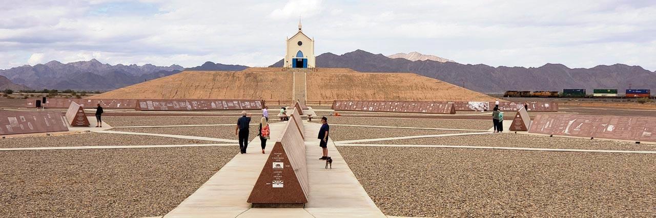 Visitors to the History of Humanity in Granite peruse the Missouri Red granite monuments on the grounds in front of the austere white Church on the Hill and mountain ranges in the background