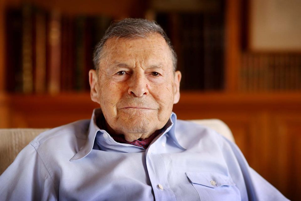 A smiling elderly man in a blue collared shirt and red patterned neckerchief