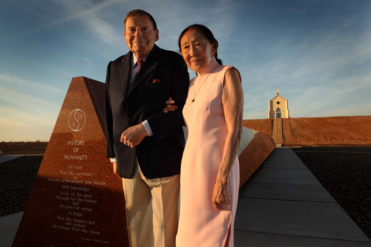 Felicia wearing a pink sleeveless high-collared dress gently holding Jacques arm, an elderly man in black sports coat, slacks, and tie, in front of a History of Humanity monument