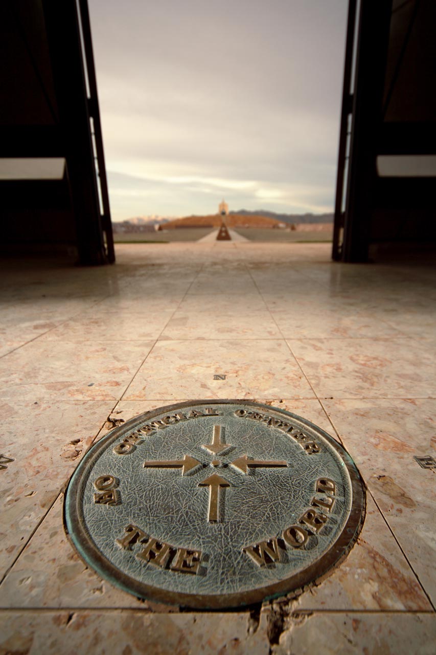 Metal plaque on a marble floor which reads Official Center of the World with the cardinal directions around it and the Church on the Hill faintly visible in the background