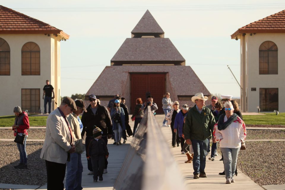 Crowd of visitors walk along granite monument with Center of the World pyramid in the background