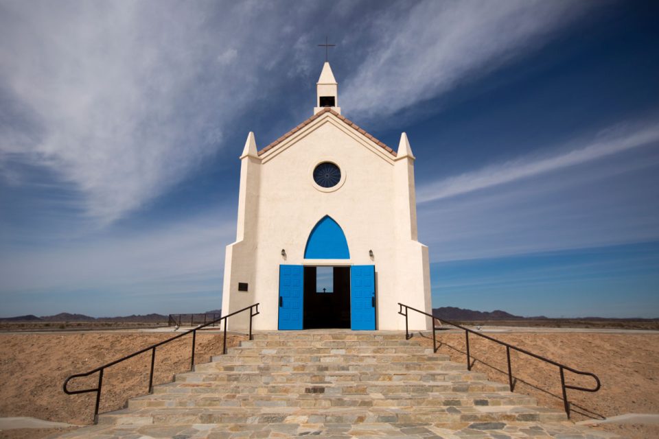 A small white church with blue doors and a single round stained glass window known as The Church on the Hill in Felicity California