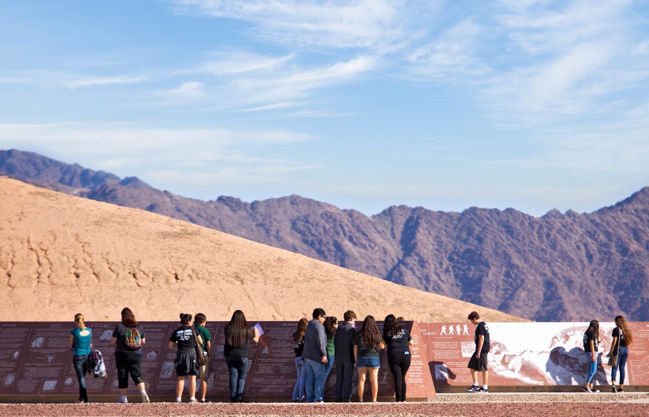 Group of young adults stand in front of granite panel at the museum of history in granite near Yuma, Arizona with mountain range in the background