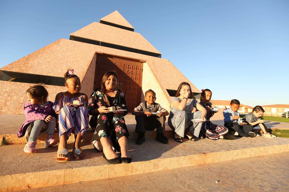 Smiling children wearing dress clothes sit in front of the center of the world pyramid eating cake