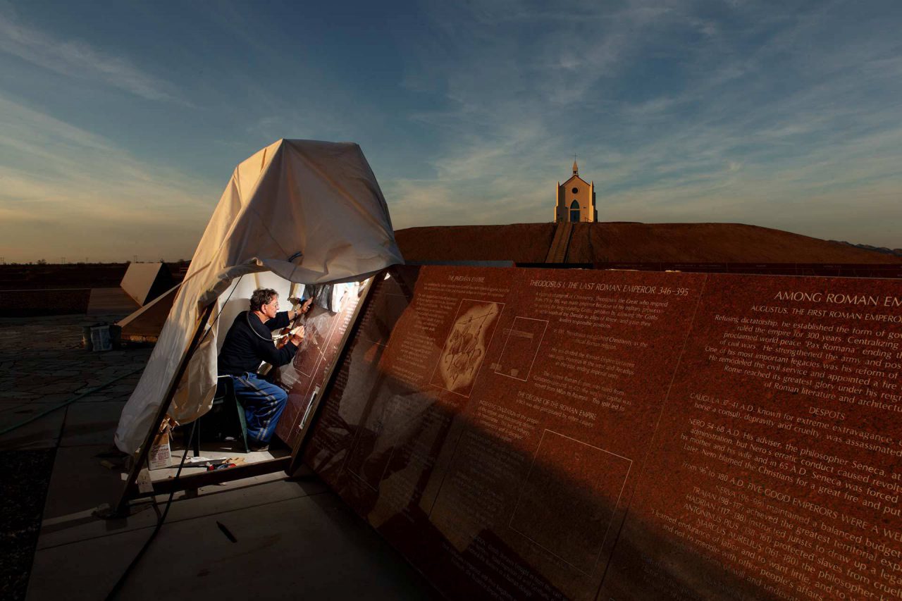 Artist engraving a granite panel beneath a tarp covering at dusk with the hill of prayer and church on the hill Felicity in the background