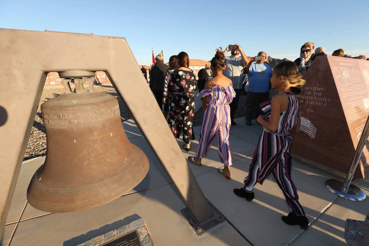 Children walking in a line between a replica of the Liberty Bell and the History of Humanity in Granite panel depicting the History of the United States of America as onlookers take pohtos