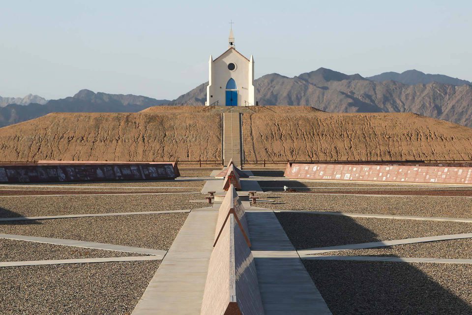 White church with blue doors and single round stained window atop the hill of prayer with History of Humanity in Granite exhibits in the foreground