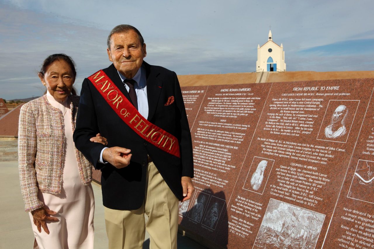 Jacques Istel wearing a sportcoat and sash which says Mayor of Felicity, arm in arm with his wife Felicity wearing a pink dress and matching coat in front of History of Humanity in Granite monument