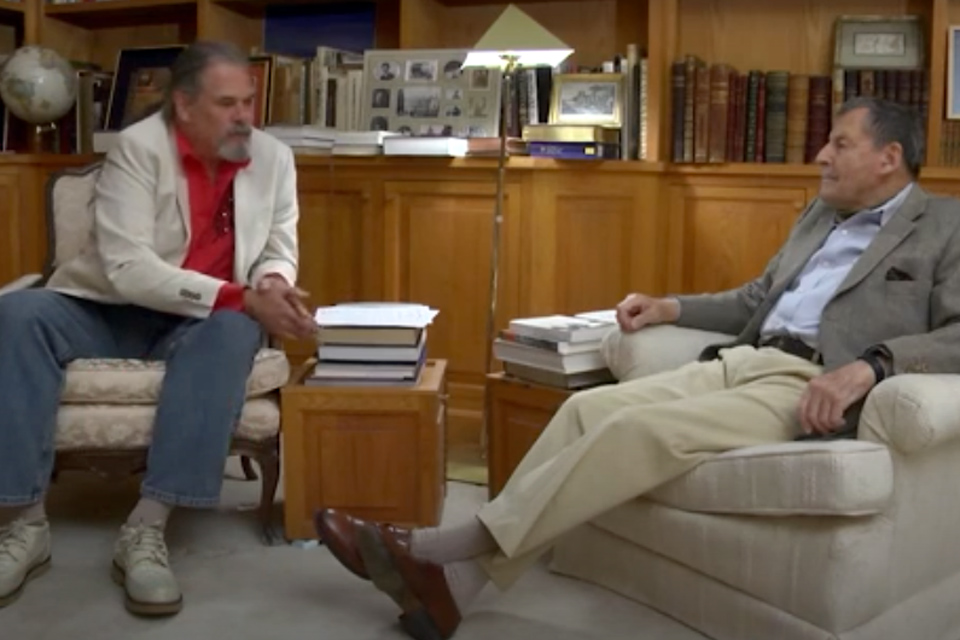 Jacques reclines in a cushioned chair with his legs extended and crossed at the ankle as he is interviewed by a man wearing a red shirt and sports coat
