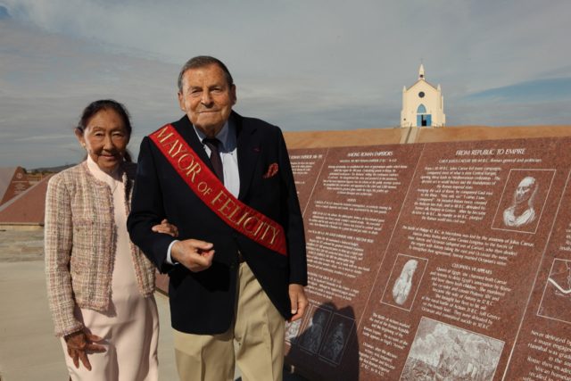 Jacques Istel wearing a sportcoat and sash which says Mayor of Felicity, arm in arm with his wife Felicity wearing a pink dress and matching coat in front of History of Humanity in Granite monument