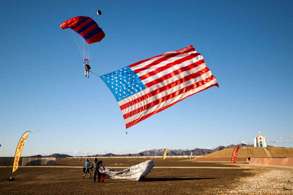 A skydiver flying beneath a red and blue parachute with the American Flag trailing below him sets up for landing at the History of Humanity in Granite as another skydiver looks on from the ground