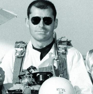 A young Jacques Istel wearing dark aviator glasses and a skydiving rig holding a helmet